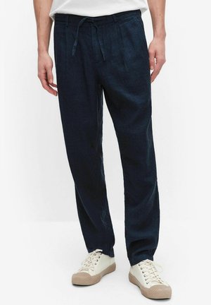 Model draagt marineblauwe casual linnen broek met tailleband met trekkoord en witte sneakers met beige zolen op witte achtergrond.