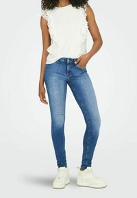 Weißes, strukturiertes ärmelloses Oberteil mit gerafften Kanten, kombiniert mit blauen Skinny-Jeans und weißen Sneakers, vor einem schlichten Hintergrund stehend.