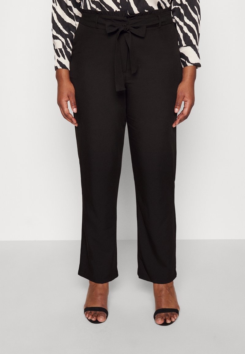 Femme portant un pantalon noir taille haute avec un détail lien à l'avant, associé à un top manches longues imprimé zèbre et des talons ouverts noirs.