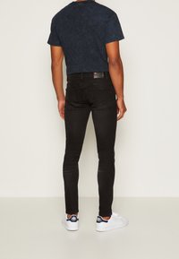 Man i svarta slim fit jeans, en marinblå kortärmad skjorta och vita Adidas Stan Smith-sneakers, vänd bort från kameran mot en enfärgad bakgrund.