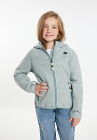 Ljusgrön zip-up fleecejacka med huva, som har en svart paraplylogotyp, texturerat tyg och dekorativa dragkedjor. I kombination med blå jeans.