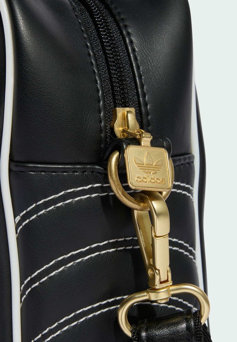 Section de sac en cuir noir avec surpiqûres blanches, fermeture éclair noire et fermoir métallique doré avec le logo Adidas et le symbole trèfle.