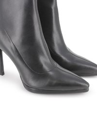 Botas de tobillo de cuero negro de tacón alto con punta afilada y costuras visibles, mostradas una al lado de la otra sobre un fondo blanco.