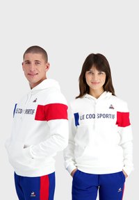Sweatshirts blancs avec des rayures rouges et bleues sur la poitrine, arborant le logo "LE COQ SPORTIF". Poches kangourou et poignets côtelés.