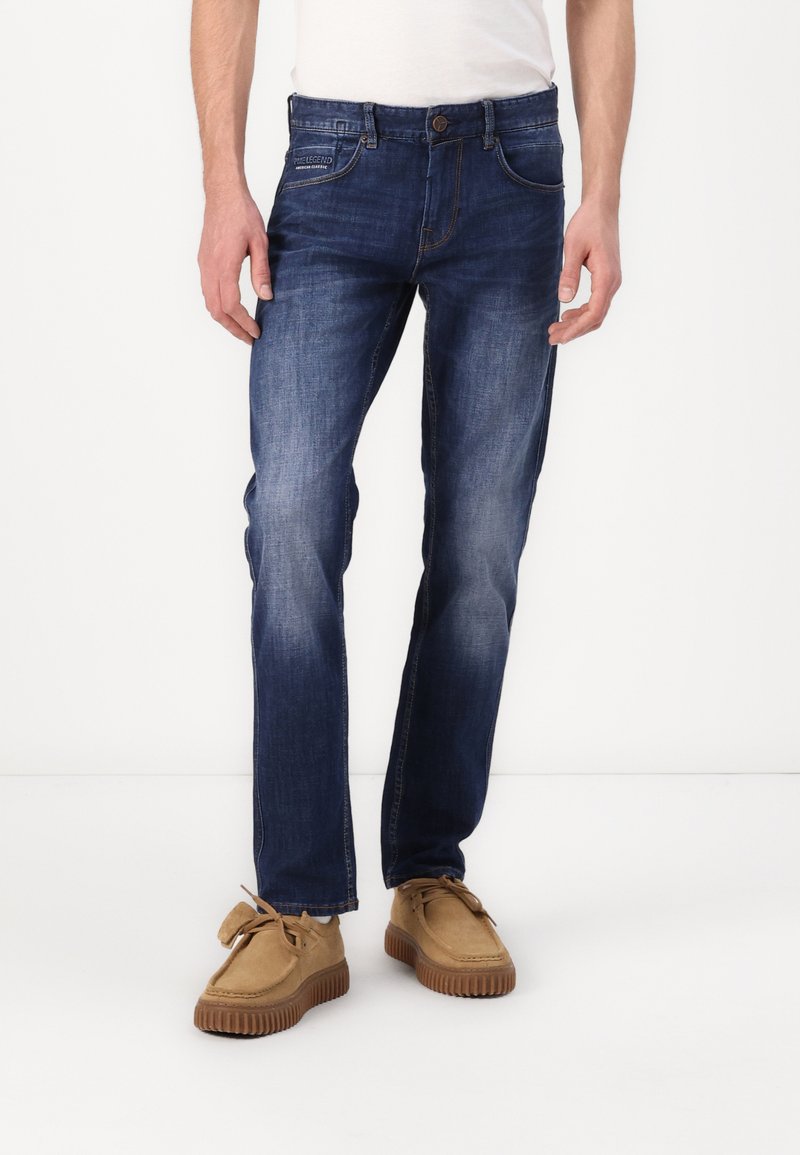 Jeans de mezclilla azul oscuro con un diseño de pierna recta. Con cierre de botón y un sutil desgastado. Llevados con zapatos de ante marrón claro.