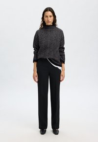 Strickpullover in Schwarz mit Fischgrätenmuster, hohem Kragen und legerem Schnitt, kombiniert mit hochgeschnittenen schwarzen Hosen und schwarzen Schuhen.