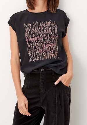 Kobieta nosząca czarny top bez rękawów z różowym abstrakcyjnym nadrukiem i napisem, połączony z czarnymi spodniami z wysokim stanem, jedna ręka w kieszeni.