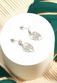 Silverfärgade metallörhängen med en utskuren bladdesign, med en polerad yta och platt bakhuvud, visade på en cirkulär vit pedestal.