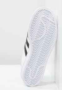 Vit sportskor med svarta ränder, med en texturerad gummisula. Skons sida visar en slät läderöverdel och detalj med varumärke.