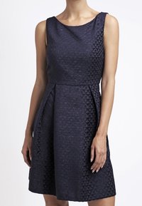 Granatowa sukienka bez rękawów z teksturowanym, geometrycznym wzorem, dopasowanym korpusem i rozkloszowanym dołem, sięgającym tuż ponad kolano.