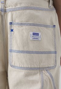 Pantalones de mezclilla beige con costuras azules y un bolsillo trasero. Presenta una etiqueta blanca con texto azul, que detalla la marca y la información de cuidado.