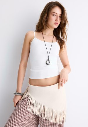 Mladá žena so bielym tielkom, nohavicami v káro, béžovou strapcovitou zavinovacou sukňou, čiernym náhrdelníkom s príveskom a náramkom so cvokmi pózuje pri bielej stene.