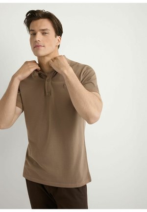 Młody mężczyzna poprawiający kołnierz brązowego polo z krótkim rękawem, stojący na tle jednolitego jasnego tła.