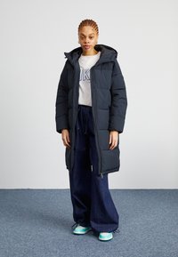 Marineblauwe puffercoat met een capuchon, voorzien van een ritssluiting aan de voorkant en elastische manchetten, gedragen over een lichtgrijs shirt en een wijde spijkerbroek.