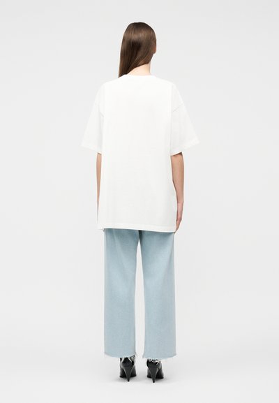 Γυναίκα με μακριά μαλλιά, ντυμένη με λευκό υπερμεγέθη κοντομάνικο πουκάμισο, ανοιχτού μπλε παντελόνι με φαρδιά πόδια και μαύρες ψηλοτάκουνες γόβες, γυρισμένη πλάτη.