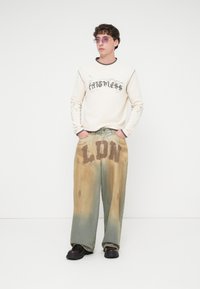 Εκρού φούτερ με μαύρο κείμενο και γραφικά· φαρδιά τζιν παντελόνια με ξεθωριασμένο πράσινο χρώμα και το "LDN" τυπωμένο καφέ στη μπροστινή πλευρά.