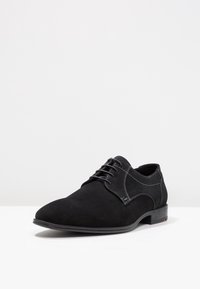 Chaussure noire en daim à lacets avec un bout pointu, coutures contrastées et semelle en caoutchouc plate. Présente une texture lisse et un design minimaliste.