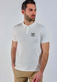 Homme portant un polo blanc avec le logo "Patrouille de France 1953", pantalon beige, se tenant avec les mains dans les poches, regardant sur le côté.