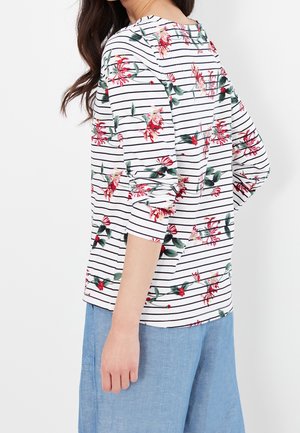 Femme portant une chemise blanche à manches longues avec des rayures horizontales noires et un imprimé floral rouge, associée à un pantalon ample bleu clair.