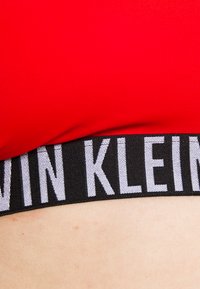 Červená elastická sportovní podprsenka s širokým černým elastickým pásem, na kterém je bílýlogo "CALVIN KLEIN". Hladká textura, přiléhavý střih.