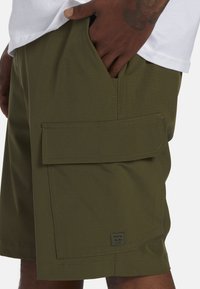 Shorts cargo verts avec une grande poche latérale, texture lisse et coutures visibles. Étiquette de logo près de l'ourlet.