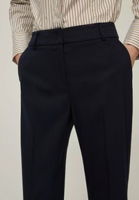 Persona vestita con pantaloni neri a vita alta e sartoriali e una camicia a righe beige e bianca a maniche lunghe con le mani infilate nelle tasche.