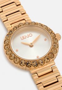 Reloj en tono dorado con una caja redonda adornada con cristales de color marrón claro, un dial de ráfaga solar y una pulsera de eslabones metálicos integrada. Nombre de la marca exhibido.