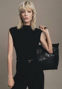Top negro sin mangas con cuello alto, combinado con un cinturón negro. La modelo sostiene un gran bolso tote de cuero negro texturizado. Fondo neutro.