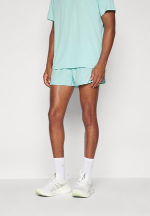 Personne portant une chemise à manches courtes bleu clair et un short assorti, des chaussettes blanches montantes et des chaussures de course blanches, se tenant devant un fond uni.