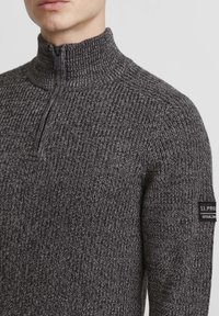 Strikket zip-sweater i mørkegrå med et tekstureret mønster, der har en høj krave og et lille mærke på venstre ærme.