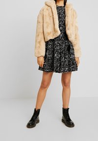 Beige fuskpäls cropped jacka över en svart blommig klänning med volangkjol, ihop med svarta snörskor. Slät textur och avslappnad design.