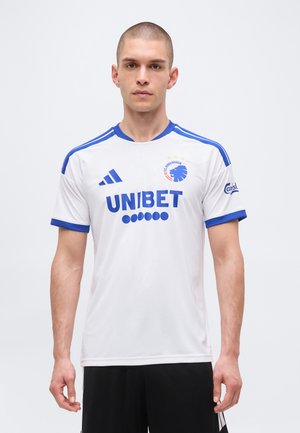 Mand iført hvid og blå FC København fodboldtrøje med UNIBET og Carlsberg logoer, står foran en ensfarvet baggrund.
