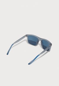 Ochelari de soare cu nuanță albastră, având o ramă gri mată. Designul prezintă laturi drepte și o formă ușor unghiulară. Accentele includ vârfuri albastre.