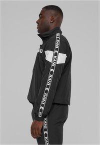 Juodas vėjo nepraleidžiantis švarkas su aukšta apykakle, su baltais akcento juostelėmis ir prekės ženklu ant rankovių; lygus, lengvas audinys; elastiniai rankogaliai.