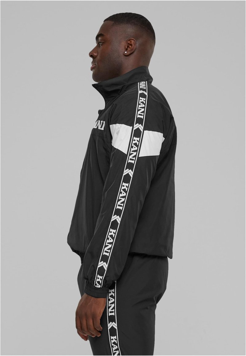 Juodas vėjo nepraleidžiantis švarkas su aukšta apykakle, su baltais akcento juostelėmis ir prekės ženklu ant rankovių; lygus, lengvas audinys; elastiniai rankogaliai.