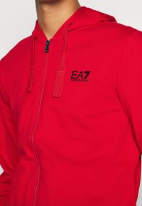 Sudadera con capucha roja con cremallera, acabado texturizado, con logo EA7 negro, capucha ajustable con cordón y cremallera en contraste.