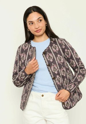Frau trägt eine gesteppte dunkle Jacke mit Blumenmustern über einem hellblauen Top und weißen Hosen und hält mit einer Hand den Kragen der Jacke.