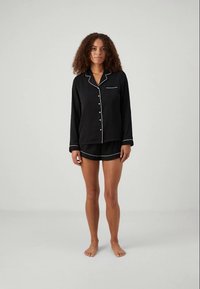 Kvinna som står barfota iklädd en svart knapp-up långärmad pyjamasöverdel och matchande shorts med vit piping mot en enfärgad bakgrund.