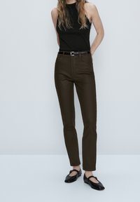 Vrouw in een mouwloze zwarte top, donkerbruine high-waist slim pants met riem en zwarte Mary Jane flats, staande tegen een effen achtergrond.