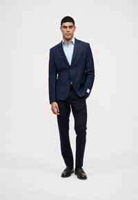 Blazer azul-marinho com um único botão, combinado com uma camisa azul clara, calças ajustadas e sapatos pretos. Tecido suave com um corte moderno.