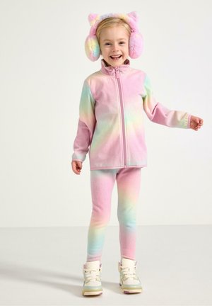 Smilende ung pige iført pastel regnbue fleecejakke og leggings med cremefarvede sneakers og bløde regnbue-ørevarmere med katteører.