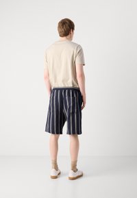 Chemise beige à manches courtes, short rayé bleu marine avec taille élastique et poches latérales, porté avec des chaussures blanches et des chaussettes beiges.