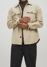 Beige wool blend shirt jacket featuring two chest pockets, black buttons, and a slightly curved hem, layered over a brown turtleneck.