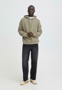 Olijfgroene capuchontrui met grijze trekkoorden, gedragen over een wit shirt. Gecombineerd met donkergrijze wijde broekspijpen en beige sneakers.