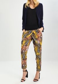 Pantalons jaunes à motifs, avec imprime de feuillage tropical, associés à un haut noir et un blazer bleu marine. Des sandales noires à talons complètent la tenue.