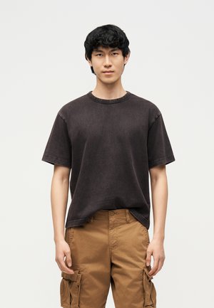 Νεαρός με μαύρα σγουρά μαλλιά, ντυμένος με ξεθωριασμένο μαύρο t-shirt και καφέ cargo σορτς, στέκεται μπροστά σε ένα απλό φωτεινό φόντο.