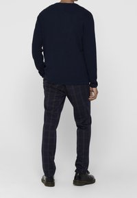 Laivastonsininen teksturoitu neulepaita, yhdistettynä tummiin ruutuhousuihin, joissa on vaaleanruskeita viivoja, täydentäen mustilla kengillä.