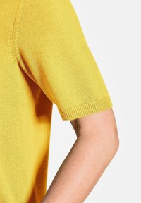 Kurzarmpullover aus gelbem Strick mit weicher Textur, gerippten Abschlüssen und lässiger Passform. Nahtdetails sorgen für subtilen Designinteresse.
