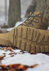 Brown hiking boots with textured soles rest on snowy ground scattered with dry leaves in a forest setting.