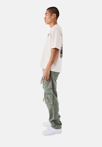 Camiseta de manga corta beige con un gráfico en la espalda, combinada con pantalones cargo verde oliva que cuentan con múltiples bolsillos y cremalleras. Zapatillas blancas.